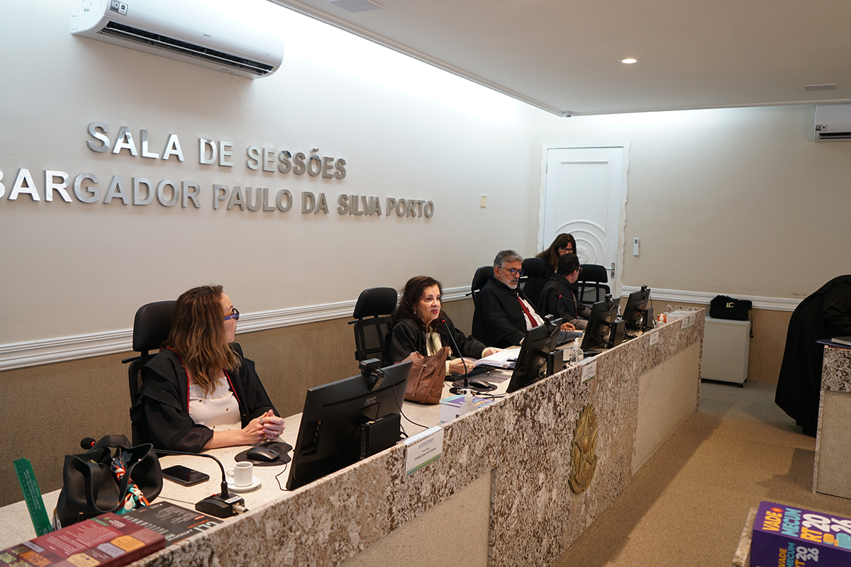 A foto mostra a mesa diretora com três magistrados de toga preta e gola vermelha; uma mulher à esquerda mexe no notebook, enquanto no centro a desembargadora Fernanda Uchôa preside o julgamento, flanqueada por um colega e por uma servidora ao fundo.