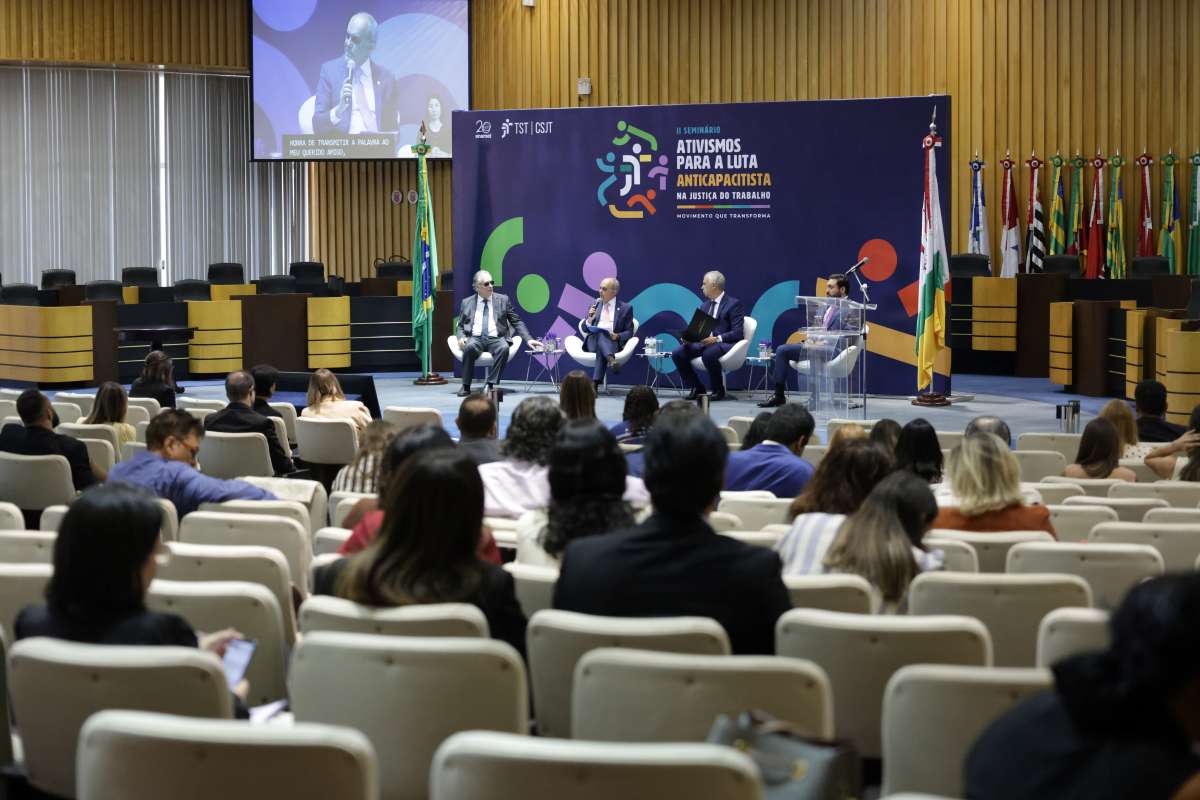Vista do público de um auditório lotado acompanhando um painel composto por quatro homens de terno sentados no palco. Ao fundo, destaca-se um grande painel roxo com detalhes coloridos e a inscrição "II SEMINÁRIO ATIVISMOS PARA A LUTA ANTICAPACITISTA NA JUSTIÇA DO TRABALHO", além das siglas do TST e CSJT. Acima, à esquerda, há uma grande tela de transmissão ao vivo com legenda, e ao lado direito do palco estão posicionadas diversas bandeiras oficiais.
