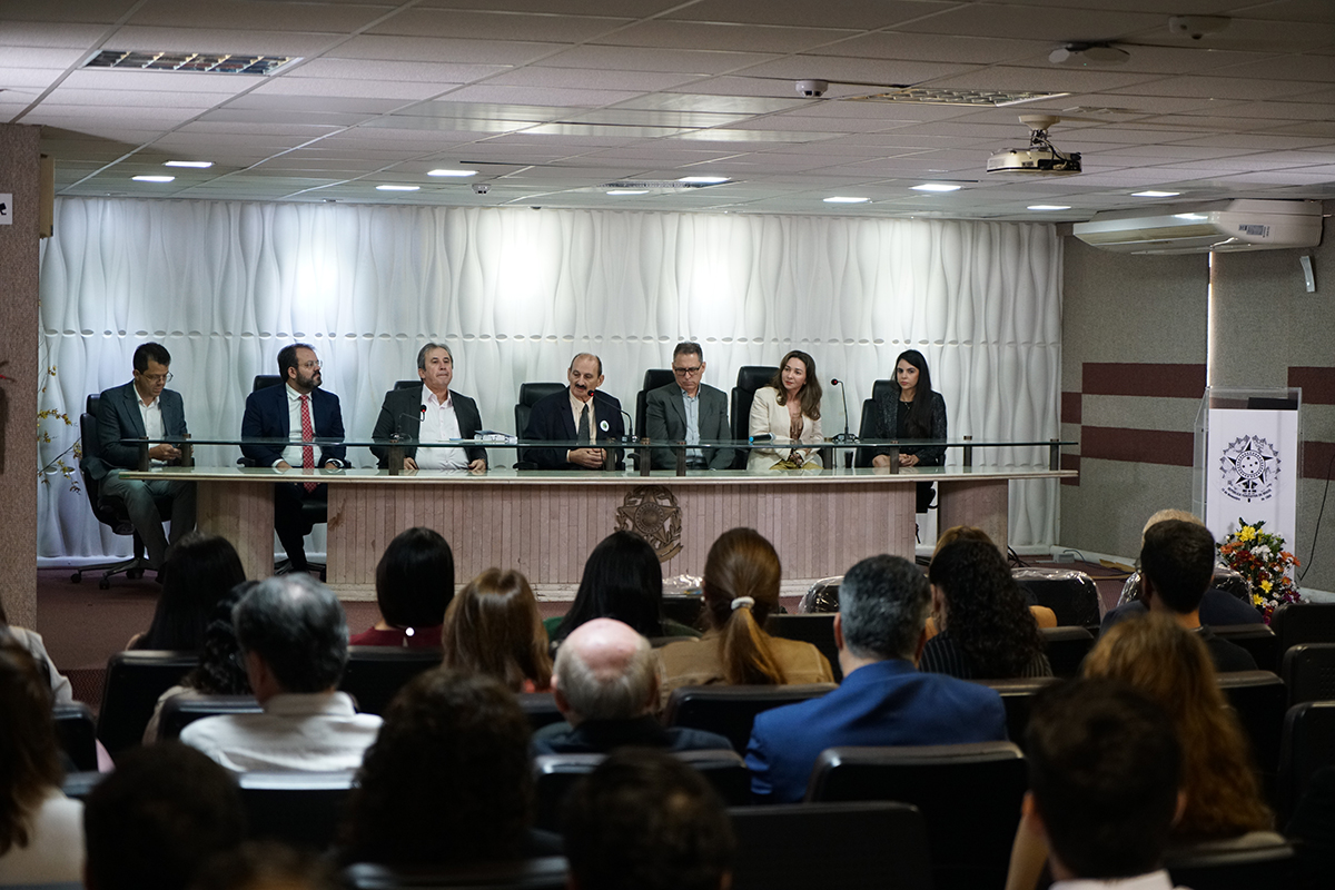 Fotografia geral do auditório mostrando a mesa de honra composta por sete autoridades sentadas durante a cerimónia. Em primeiro plano, de costas para a câmara, observa-se o público atento a ocupar os lugares do auditório. O ambiente é iluminado por focos no teto falso e decorado com arranjos florais nas extremidades do palco.
