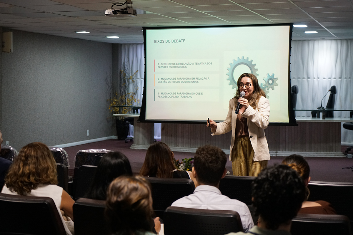A auditora-fiscal do trabalho Maria Ervanis Brito discursa ao microfone no palco diante da plateia sentada no auditório. Atrás da palestrante, a tela de projeção exibe um slide com o título "EIXOS DO DEBATE" e três pontos numerados ao lado do desenho de engrenagens. A imagem é captada a partir de trás dos participantes que assistem à palestra.