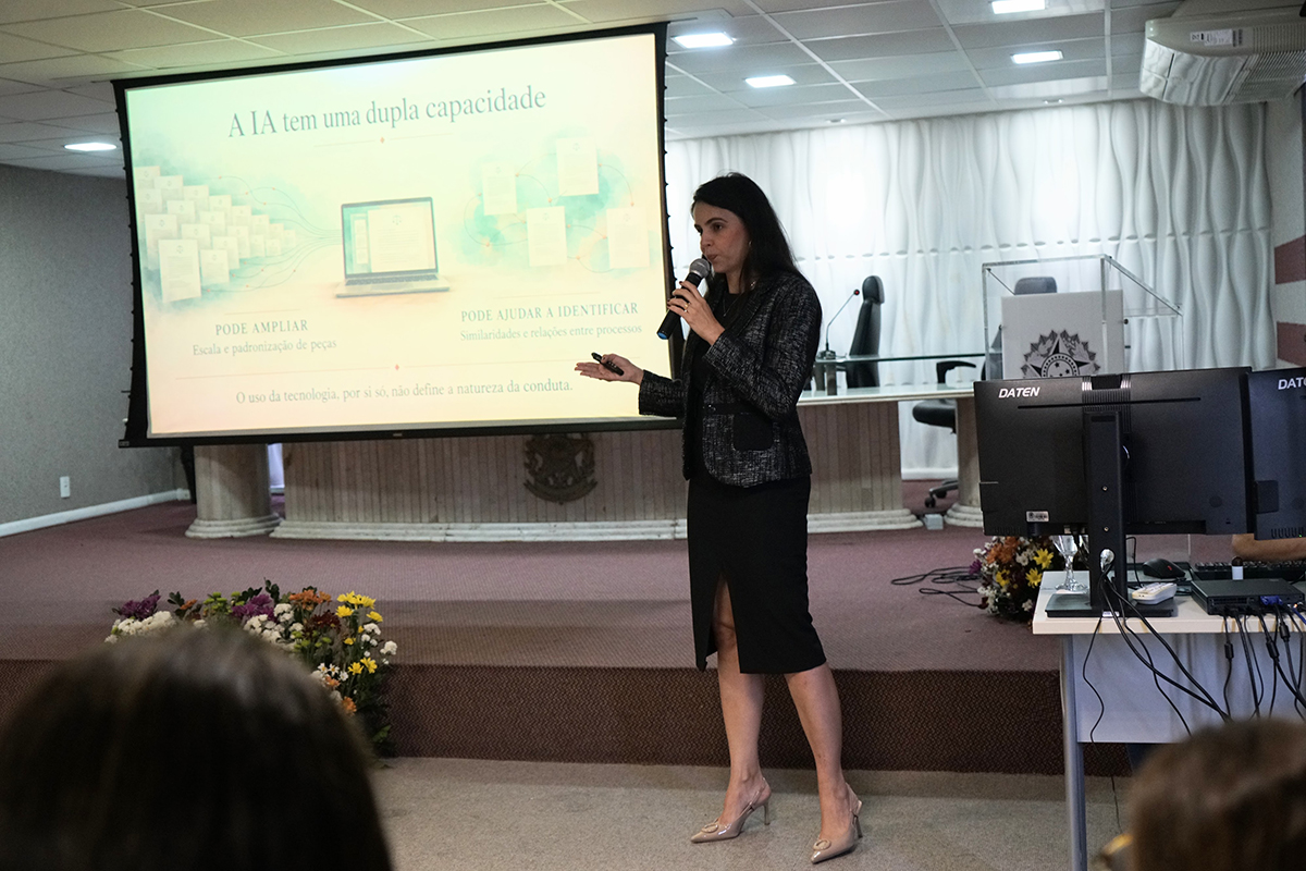 Em plano médio, a juíza do trabalho Natália Alves fala ao microfone enquanto realiza uma apresentação no auditório, posicionada ao lado de uma tela de projeção. A tela exibe um slide explicativo sobre inteligência artificial com o título "A IA tem uma dupla capacidade". Em primeiro plano, no canto inferior direito, sobressai um monitor de computador e equipamento técnico.