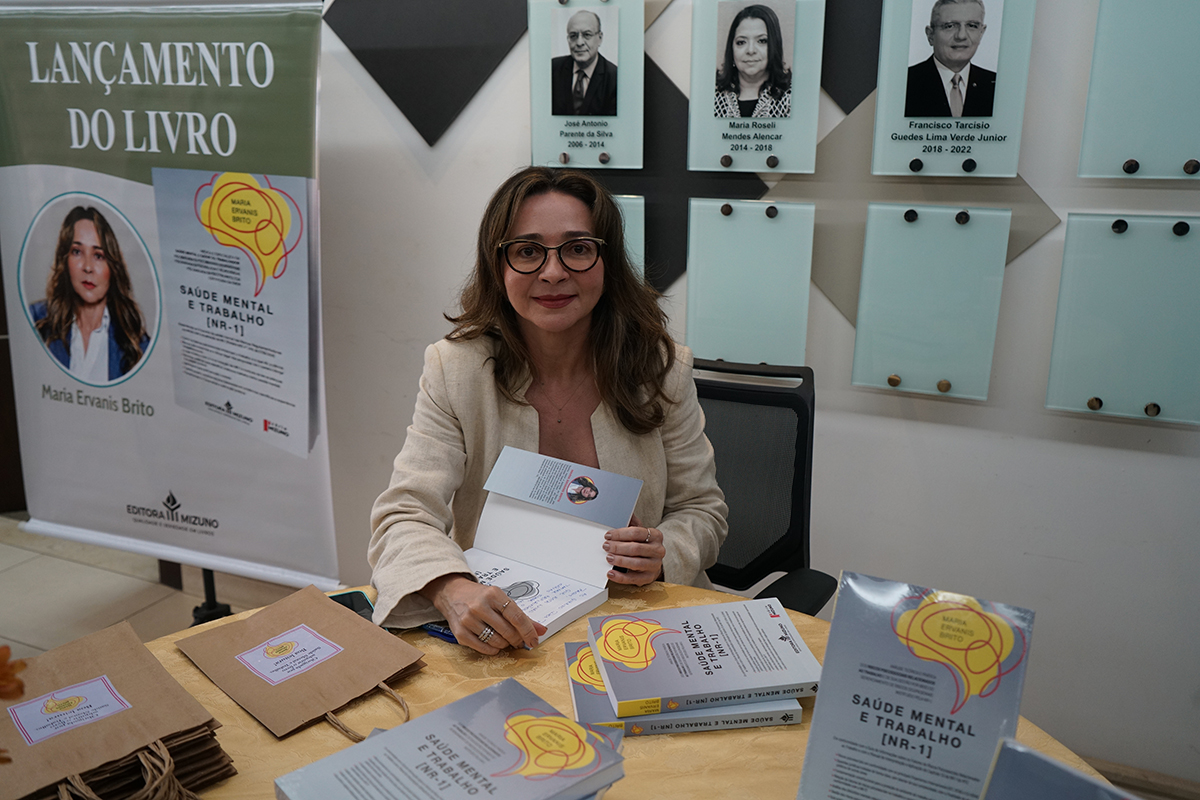 A autora Maria Ervanis Brito surge sentada à mesa durante a sessão de autógrafos, olhando para a câmara com um sorriso sereno. Sobre a mesa e à sua frente encontram-se vários exemplares do livro "Saúde Mental e Trabalho [NR-1]" e sacos de papel. Ao lado esquerdo, sobressai um banner promocional do lançamento da obra e, ao fundo, quadros na parede.