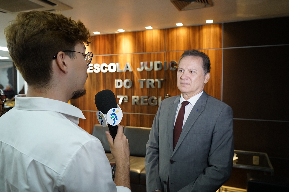Em plano médio, o superintendente regional do trabalho no Ceará (SRT-CE), Carlos Pimentel de Matos Júnior, é entrevistado por um jovem repórter que segura um microfone. O magistrado veste um fato cinzento com gravata bordô e olha com atenção para o entrevistador. Ao fundo, na parede revestida a madeira, sobressaem as letras prateadas com a inscrição "ESCOLA JUDICIAL DO TRT 7ª REGIÃO".