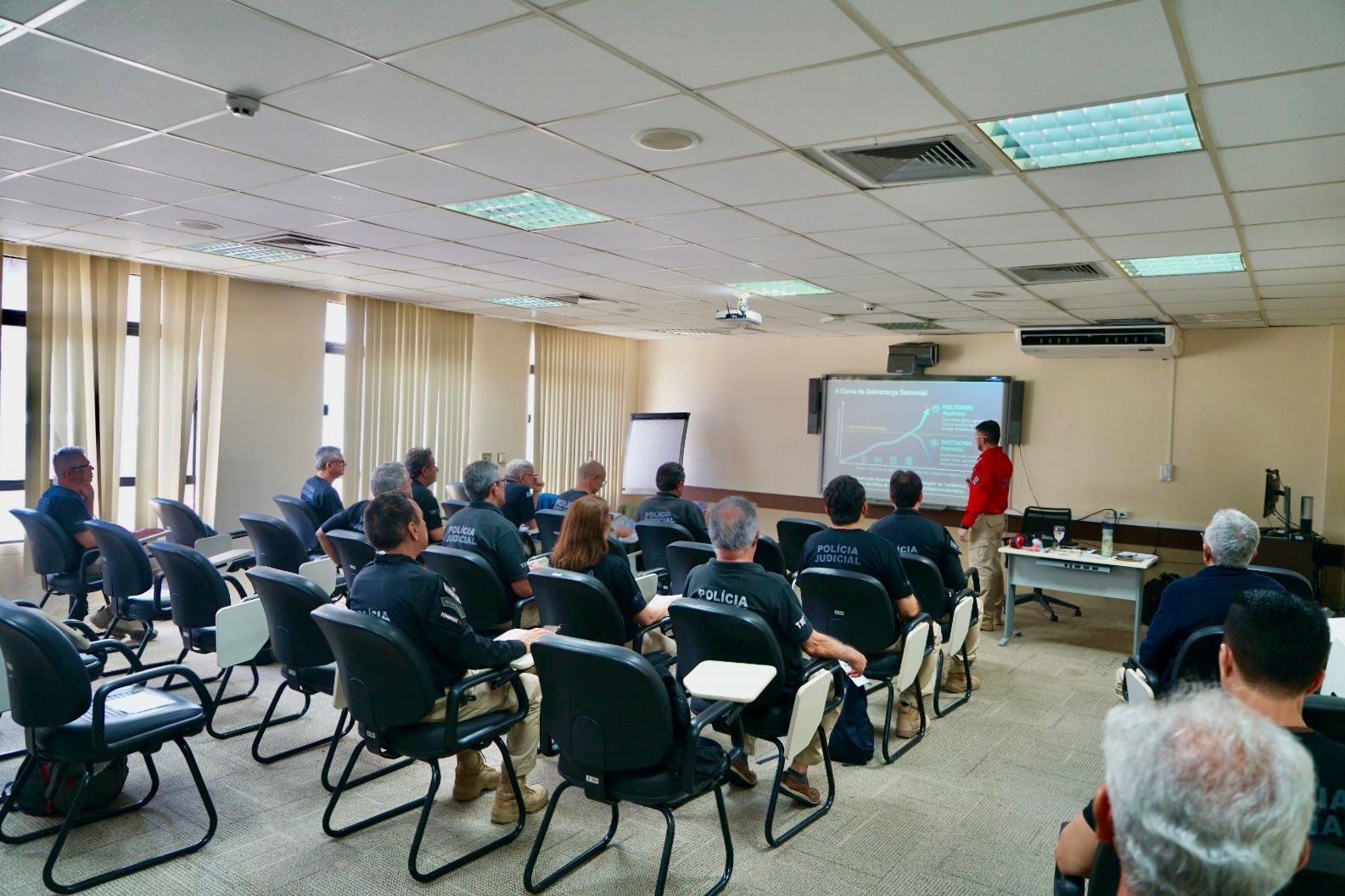 Fotografia de uma sala de treinamento, com diversos participantes sentados em cadeiras voltadas para uma tela de projeção. A maioria dos participantes veste camisas pretas com a identificação “Polícia Judicial” nas costas. À frente, um instrutor usando camisa vermelha apresenta um conteúdo projetado em uma tela, enquanto utiliza um computador sobre uma mesa. A sala possui janelas com persianas verticais, iluminação de teto, ar-condicionado e equipamentos audiovisuais. Ao fundo, há também um quadro branco.