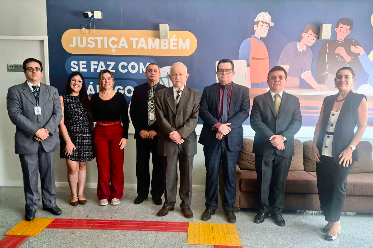 Oito pessoas estão lado a lado em pé, alinhadas para uma foto institucional. As cinco pessoas do centro e da esquerda vestem trajes formais em tons de cinza, preto e vermelho, enquanto o grupo inclui homens usando terno e gravata e mulheres em vestidos e calça social. Ao fundo, uma parede azul exibe a frase "JUSTIÇA TAMBÉM SE FAZ COM DIÁLOGO" ao lado de ilustrações de trabalhadores, com um sofá marrom posicionado à direita. No piso claro da sala, sob saem faixas de piso tátil amarelo e vermelho.