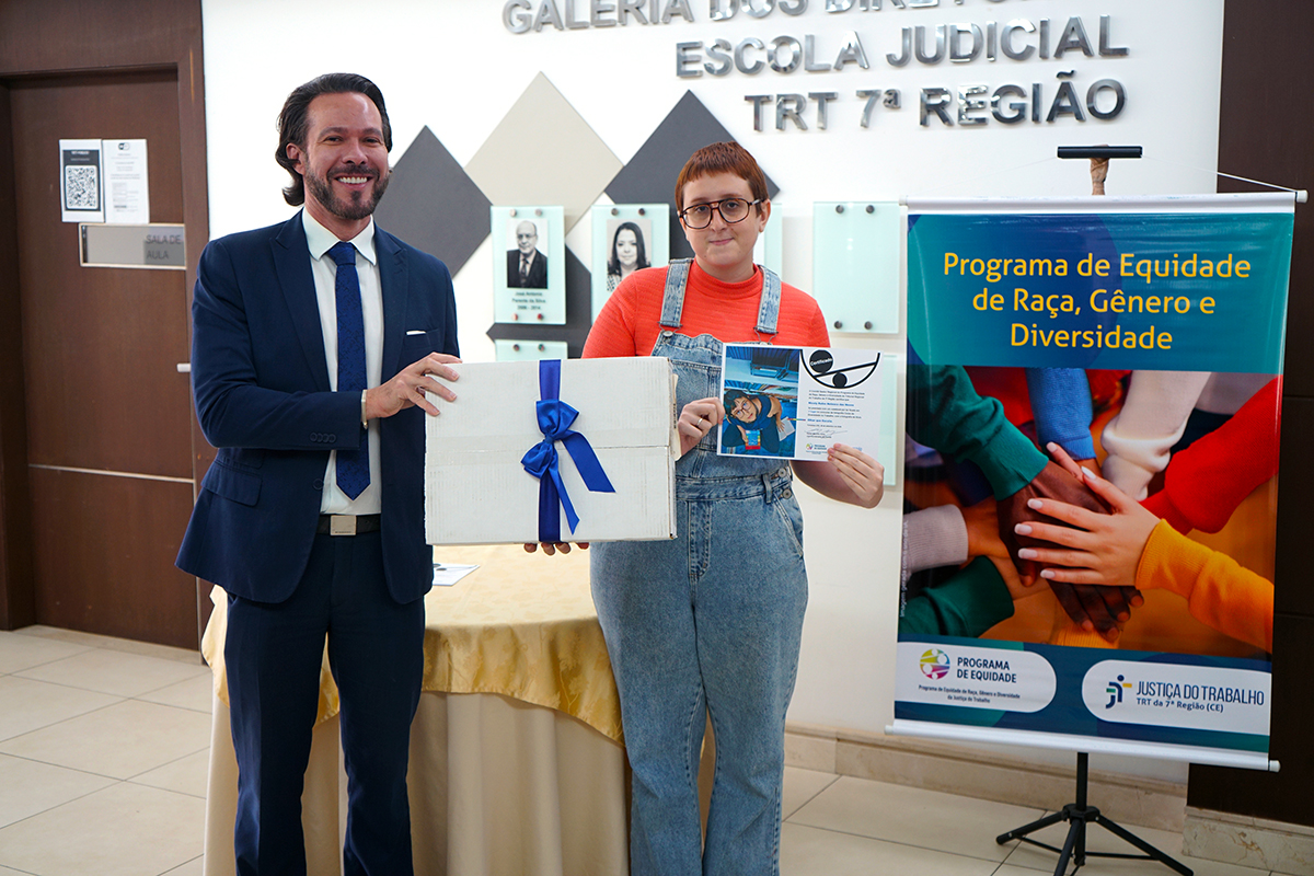 Em plano médio, um homem de terno azul-marinho entrega uma caixa branca com laço azul a uma jovem à sua esquerda. Ela vestem uma blusa alaranjada e jardineira jeans, segura o presente e exibe um certificado. Ao fundo, vê-se parte do banner do programa de equidade do TRT 7ª Região e a galeria de quadros na parede branca.