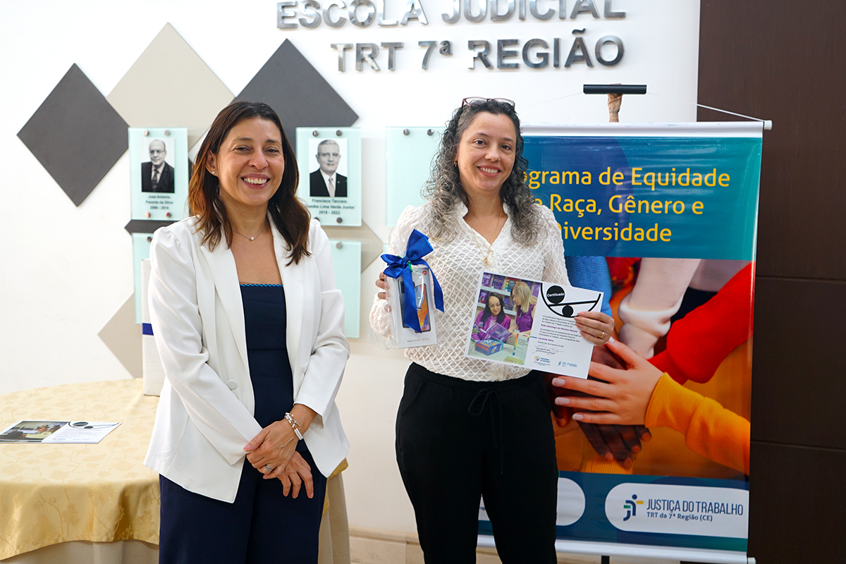 Duas mulheres sorriem lado a lado em frente à parede com a inscrição da Escola Judicial. À esquerda, uma mulher vestindo blazer branco e blusa escura está com as mãos cruzadas à frente. À direita, a outra mulher segura um certificado e um presente decorado com laço azul, enquanto o banner institucional permanece visível ao fundo.