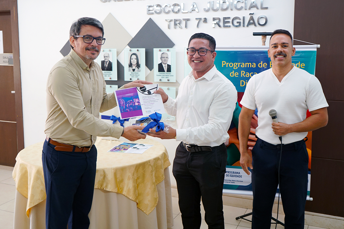 Três homens em um ambiente institucional. À esquerda, um homem de camisa bege e óculos entrega um certificado e um presente a um homem de camisa branca e óculos, no centro. À direita, um terceiro homem de blusa branca justa e calça escura segura um microfone e sorri para a câmera, com uma mesa coberta por toalha amarela e o banner ao fundo.