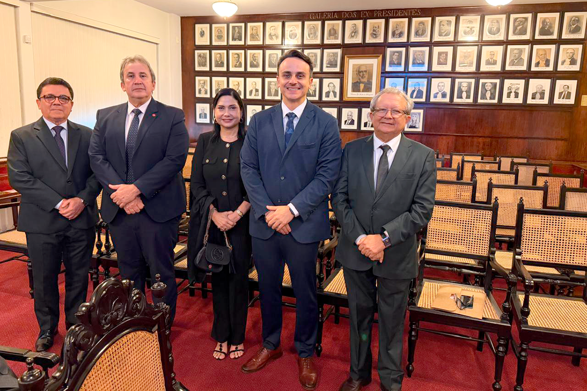 Cinco pessoas — quatro homens e uma mulher — estão em pé, lado a lado, sorrindo para a câmera em uma sala formal com carpete vermelho. Os homens vestem ternos e gravatas, e a mulher veste um conjunto preto elegante. Ao fundo, uma parede de madeira exibe diversos quadros com retratos alinhados sob a inscrição "Galeria dos Ex-Presidentes". A sala também possui cadeiras de madeira com encosto de palha dispostas ao longo do ambiente.  Audiodescrição da segunda imagem:
