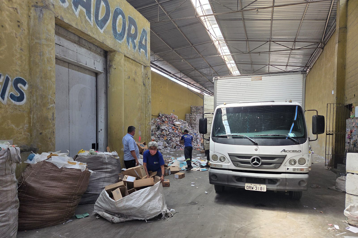 No centro da imagem, uma grande pilha de caixas de papelão e pastas de arquivos marrons e azuis está espalhada pelo chão de concreto do galpão. Na traseira aberta de um caminhão, trabalhadores entregam caixas uns aos outros, enquanto outros funcionários observam ao lado