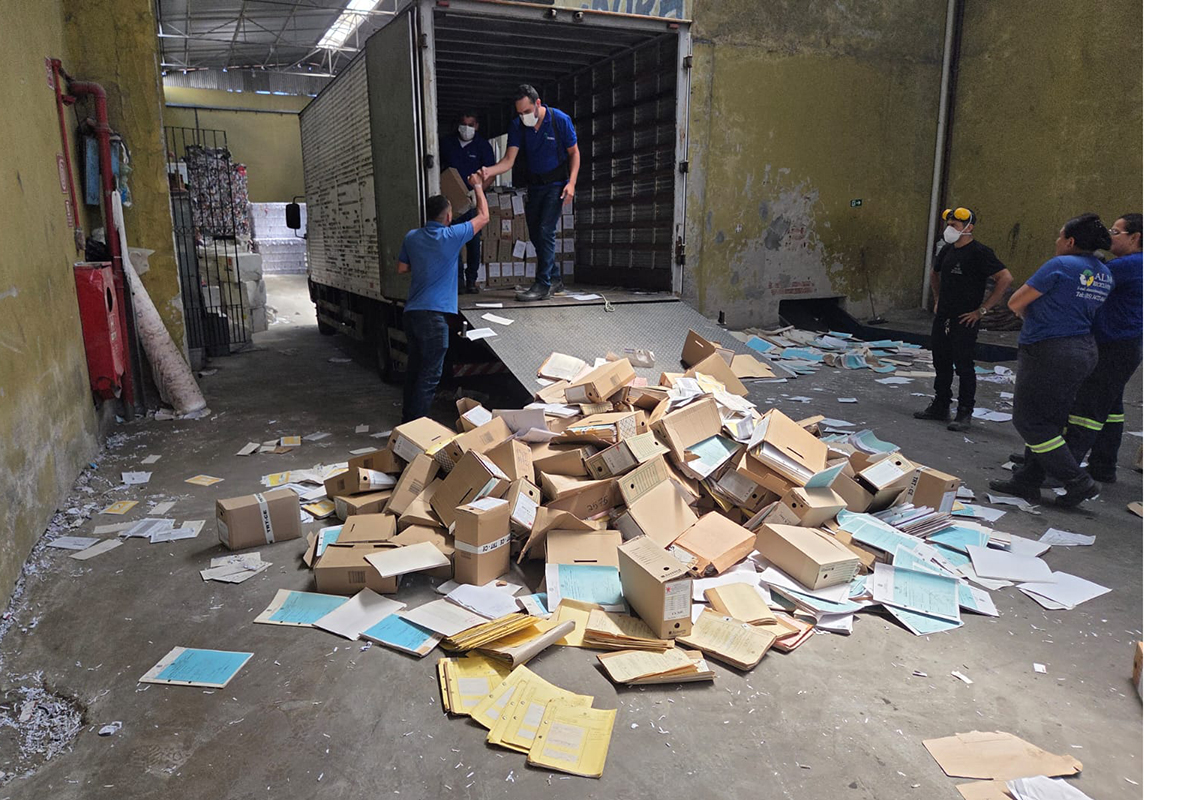 Em um galpão industrial, um caminhão baú branco da Mercedes-Benz está estacionado à direita, ao lado de colunas amarelas desbotadas. À esquerda e ao fundo, trabalhadores com uniformes azuis e máscaras organizam grandes sacos e caixas de papelão em meio a pilhas de papel impresso prensado.