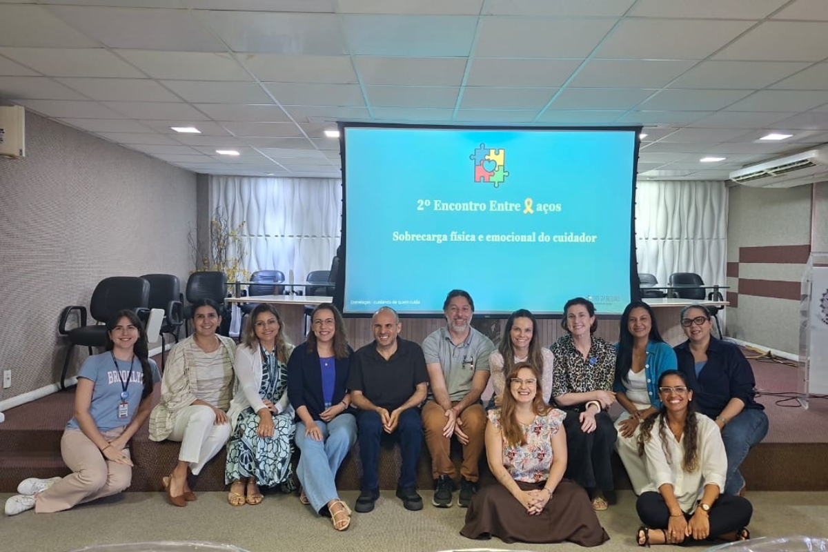 Fotografia de um grupo de 12 pessoas, entre homens e mulheres, posando sorridentes em frente a uma tela de projeção em uma sala de eventos. Na tela, aparece a identificação “2º Encontro Entre Laços”, com o tema “Sobrecarga física e emocional do cuidador” e um símbolo colorido de peças de quebra-cabeça. Ao fundo, há cadeiras, cortinas claras e uma mesa, compondo o ambiente de realização do encontro.