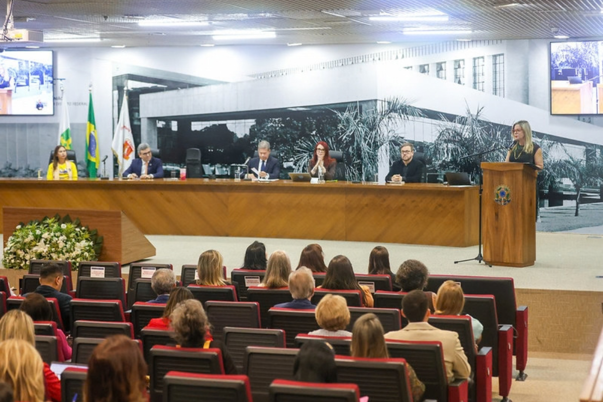 Fotografia de um evento institucional realizado em um auditório, com seis pessoas compondo a mesa de autoridades e uma mulher falando ao púlpito à direita. Ao fundo, há bandeiras e telas de exibição. O público está sentado em fileiras de cadeiras voltadas para o palco, acompanhando a programação.