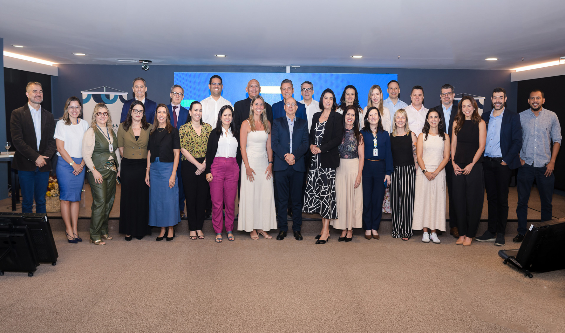 Fotografia de grupo com aproximadamente 30 pessoas, entre homens e mulheres, posicionadas lado a lado em um ambiente interno, durante um evento institucional. Os participantes vestem trajes sociais e esporte fino, em diferentes combinações de cores e estilos, e posam sorrindo para a câmera. Ao fundo, há um painel digital azul, paredes escuras e estruturas de apoio à apresentação. O espaço possui iluminação artificial, piso acarpetado e equipamentos audiovisuais nas laterais, compondo o cenário de um encontro profissional.