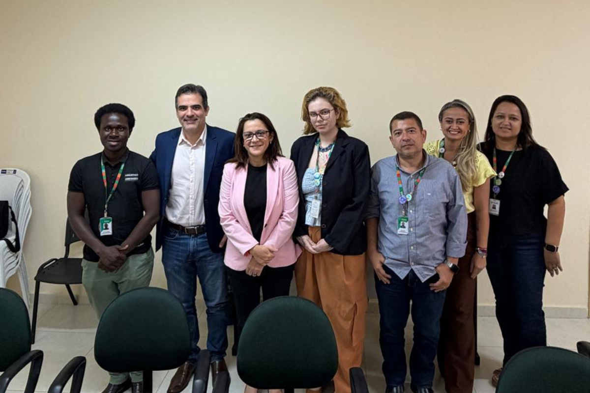 Fotografia de grupo com oito pessoas, sendo cinco mulheres e três homens, posando lado a lado em uma sala com paredes claras. Os participantes vestem roupas casuais e sociais e, em sua maioria, utilizam crachás de identificação pendurados no pescoço. Ao fundo, há cadeiras dispostas junto à parede.