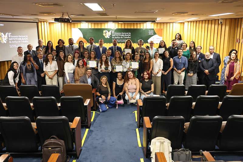Foto em plano geral de um grupo grande de pessoas reunidas no palco de um auditório, com algumas agachadas à frente. Vários participantes exibem certificados e troféus, celebrando no evento com o banner institucional ao fundo. Em primeiro plano, veem-se as fileiras de poltronas pretas do auditório com um corredor central.