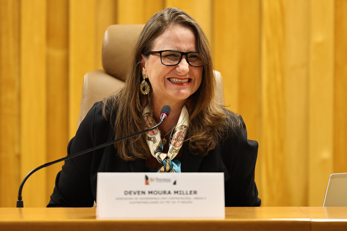 Mulher sorridente de óculos de armação preta, cabelos castanhos claros e lenço estampado no pescoço fala ao microfone. Ela vestida com blazer escuro está sentada atrás de uma bancada de madeira com uma placa de identificação com o nome "DEVEN MOURA MILLER". Ao fundo, destaca-se um painel de madeira vertical.