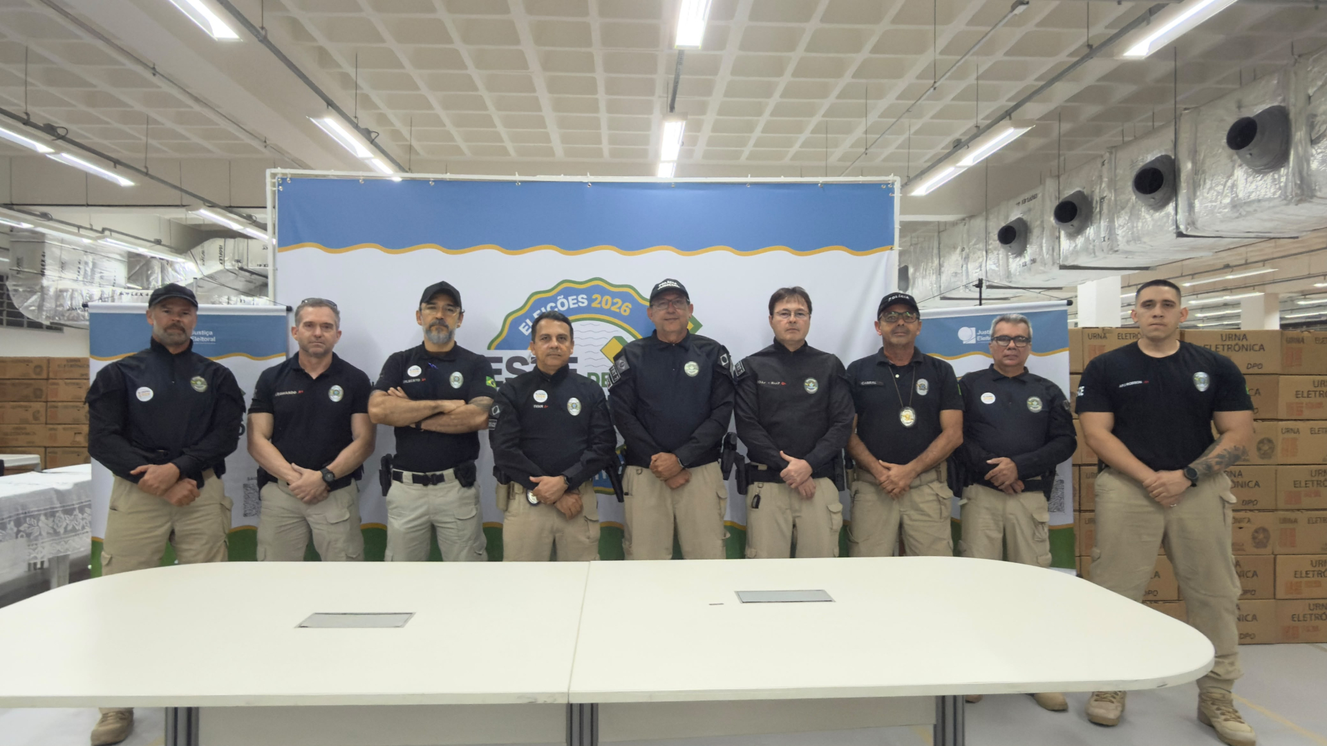 Fotografia mostra um grupo de oito profissionais de segurança, usando uniformes pretos e calças bege, posando lado a lado em um ambiente interno. Ao fundo, há um painel com a identificação “Eleições 2026” e materiais relacionados à Justiça Eleitoral. Caixas identificadas como “Urna Eletrônica” estão empilhadas nas laterais do espaço. Em primeiro plano, há uma grande mesa branca.