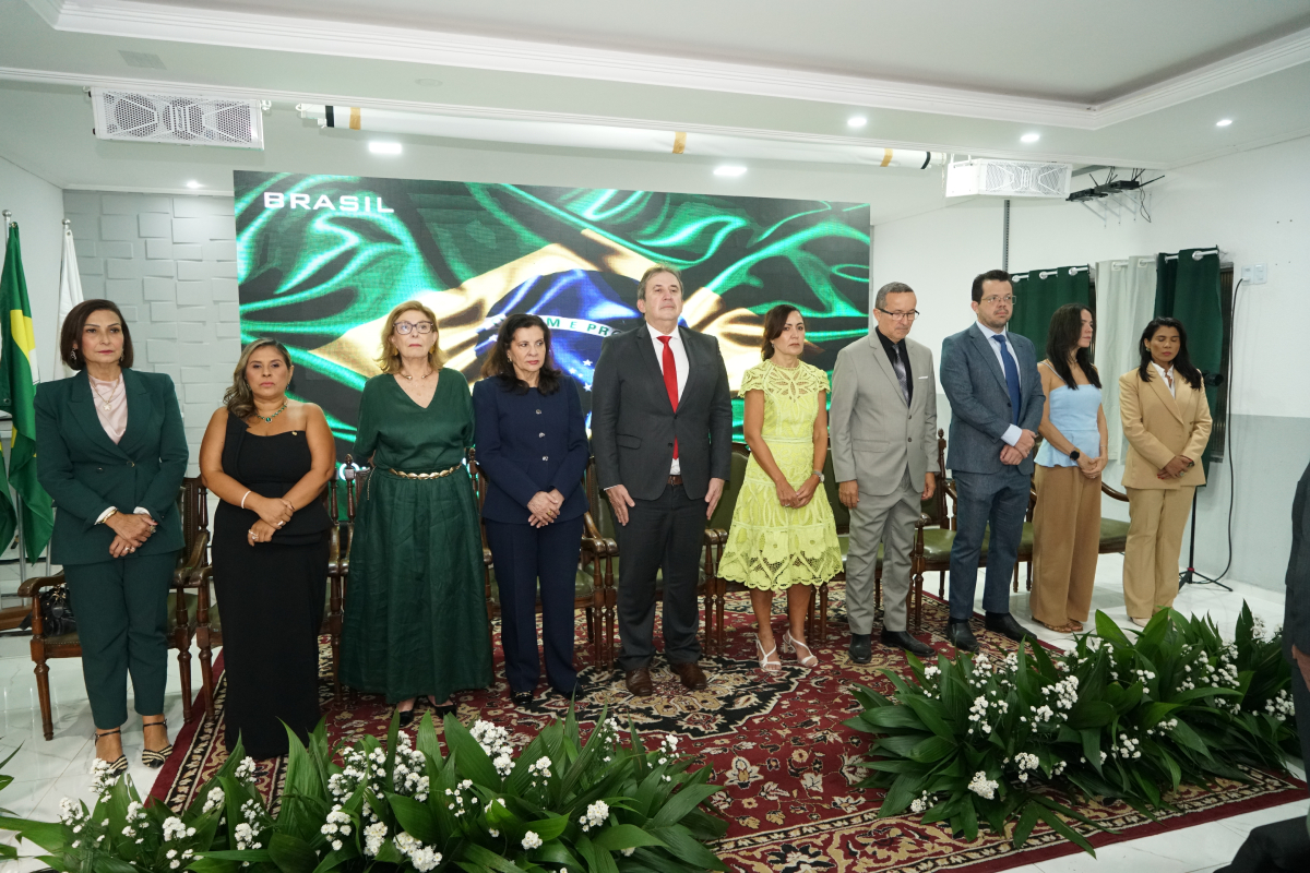 Fotografia em plano aberto e frontal de dez pessoas alinhadas lado a lado no palco de um auditório, de pé e em postura formal. Homens usam ternos e as mulheres usam vestidos ou terninhos formais em tons de verde, preto, azul, bege e amarelo. Ao fundo, um painel exibe a imagem de uma bandeira do Brasil em movimento. À frente do palco, arranjos de folhagens e flores brancas ornamentam o chão sobre um tapete estampado.