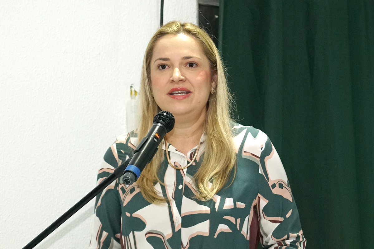 Plano médio de uma mulher de cabelos loiros e compridos vestindo uma blusa estampada em tons de verde, branco e rosa. Ela está sorrindo levemente e fala em um microfone posicionado à sua frente. Ao fundo, vê-se uma parede branca à esquerda e uma cortina verde à direita. Ela usa correntes delicadas no pescoço e brincos.