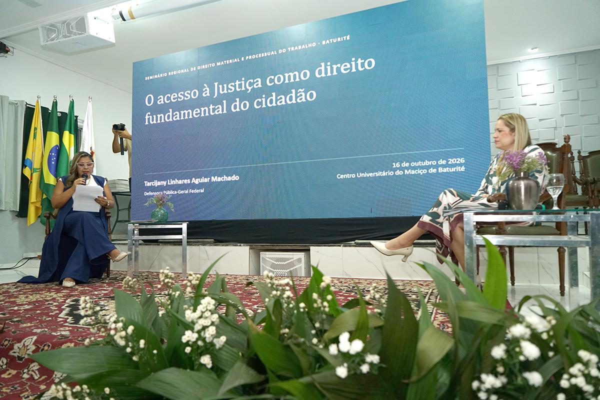 Vista do palco a partir da parte inferior, destacando arranjos de flores brancas em primeiro plano. Duas mulheres estão sentadas no palco: uma à esquerda fala ao microfone lendo um papel. Ao centro, o painel exibe o texto "O acesso à Justiça como direito fundamental do cidadão". À direita, outra mulher com vestido estampado observa atentamente.