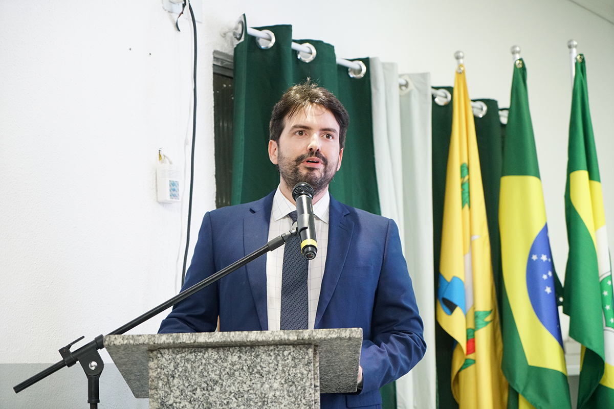 Plano médio de um homem de barba curta e cabelos escuros, vestindo paletó azul, camisa clara e gravata. Ele está posicionado atrás de um púlpito de granito e fala em um microfone fixado em pedestal. Ao fundo, veem-se cortinas verdes e bandeiras oficiais alinhadas. A parede lateral é branca e possui um interruptor visível.