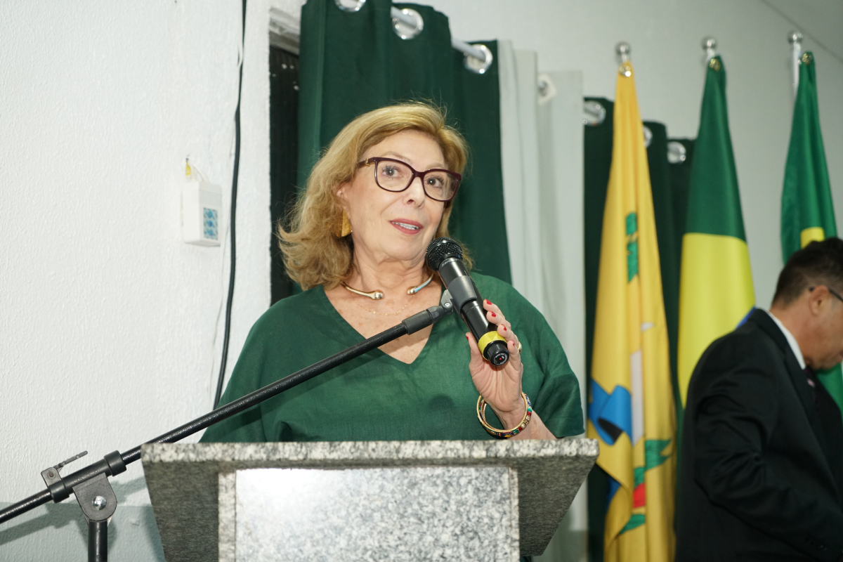 Fotografia em plano médio de uma mulher de cabelos loiros e curtos, usando óculos de armação escura e vestido verde. Ela está posicionada no púlpito de granito, segurando um microfone com a mão direita e discursando. Ao fundo, veem-se cortinas escuras e bandeiras oficiais dispostas verticalmente.