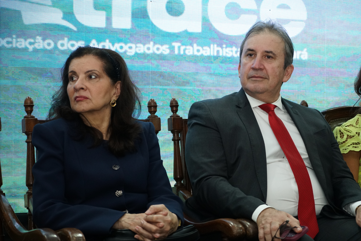 Fotografia em plano médio mostrando duas pessoas sentadas em cadeiras de madeira de estilo clássico. À esquerda, uma mulher de terno azul-marinho e cabelos escuros olha para o lado. À direita, um homem de terno cinza-escuro, camisa branca e gravata vermelha olha para a frente. Ao fundo, há um painel luminoso com textos do evento.