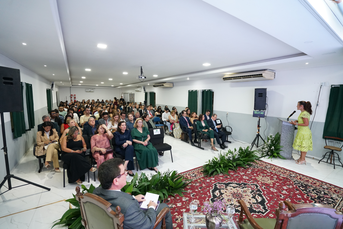 Fotografia em plano geral e elevado que mostra o auditório repleto de pessoas sentadas em fileiras de cadeiras. No canto direito, uma mulher com vestido amarelo fala em um púlpito. O ambiente é iluminado, com teto branco, piso claro e um tapete estampado avermelhado no centro, rodeado por arranjos de flores e plantas baixas.