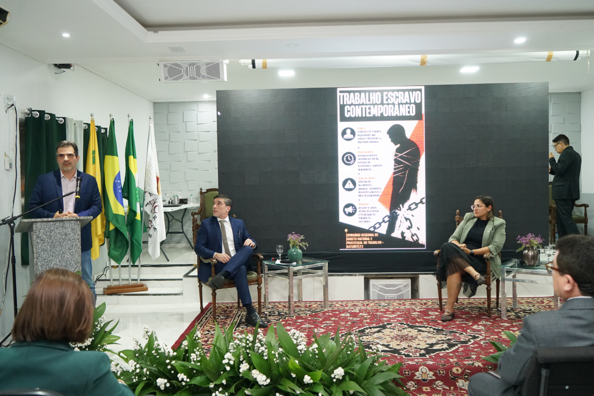 Fotografia em plano aberto mostrando um painel ao centro com a imagem de uma pessoa acorrentada e o título "TRABALHO ESCRAVO CONTEMPORÂNEO". À esquerda, um homem de paletó azul discursa em um púlpito. Outras duas pessoas estão sentadas em poltronas no palco acompanhando a apresentação. Ao fundo, estão hasteadas as bandeiras oficiais.