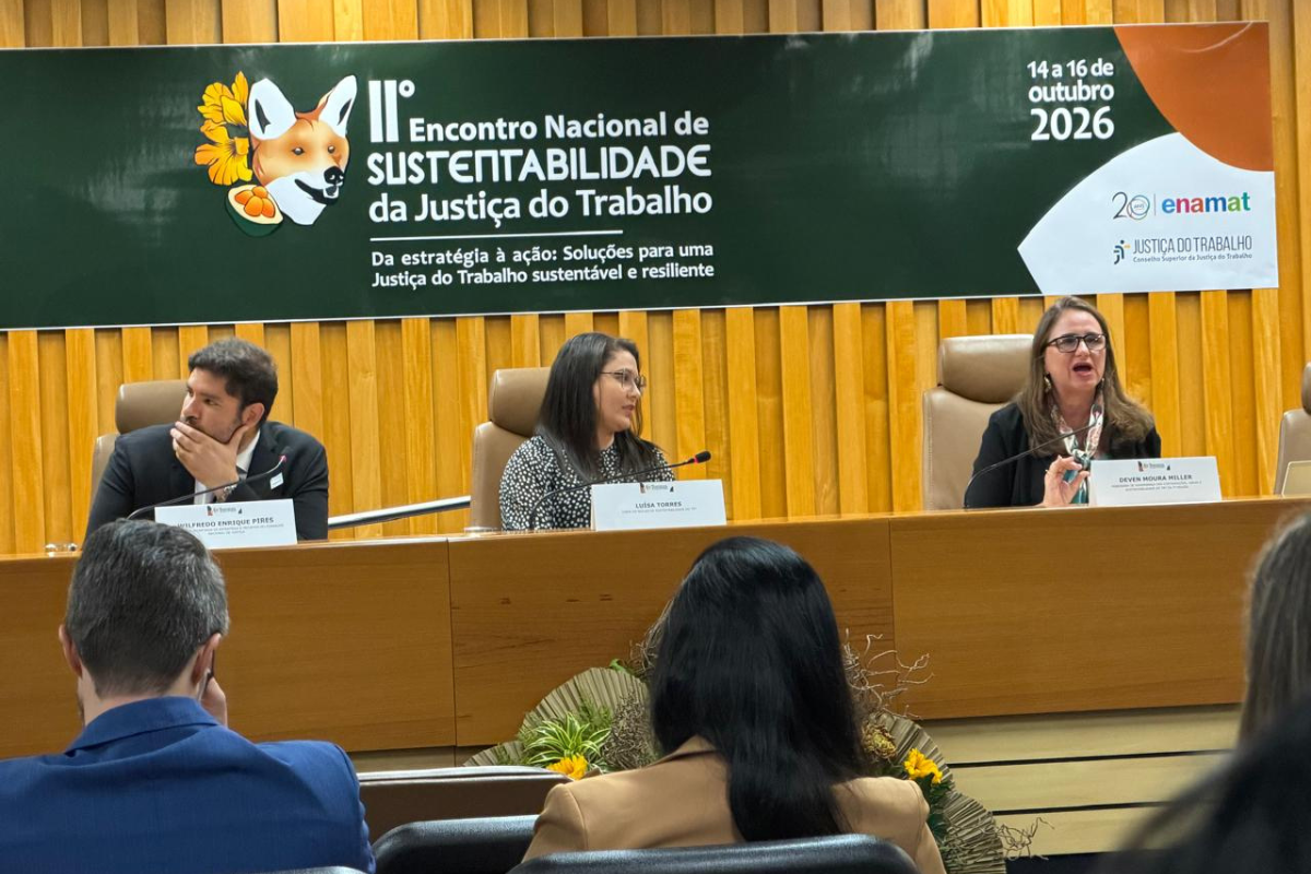 Fotografia de um painel do 11º Encontro Nacional de Sustentabilidade da Justiça do Trabalho, realizado de 14 a 16 de outubro de 2026. À frente, três participantes estão sentados à mesa de debates, com microfones e placas de identificação. Ao fundo, há um banner do evento e uma parede revestida de madeira. Em primeiro plano, pessoas acompanham a programação sentadas de costas para a câmera.