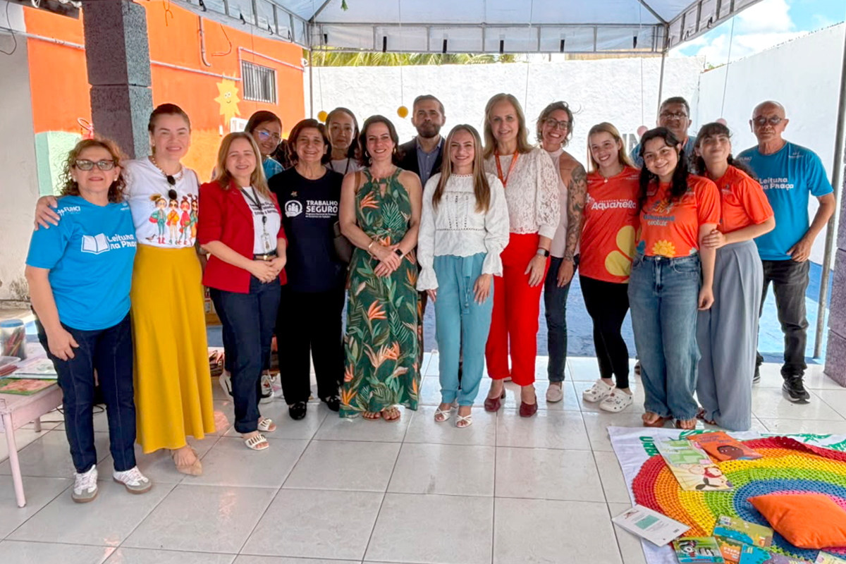 Um grupo de cerca de 13 adultos está posado lado a lado sob um toldo externo, sorrindo diretamente para a câmera. Alguns vestem camisetas de projetos sociais com frases como "Leitura na Praça" e "Trabalho Seguro". No chão, à direita, há um tapete colorido com livros infantis espalhados. A mensagem transmite um sentido de união, colaboração e integração de equipes em uma ação comunitária ou educacional.