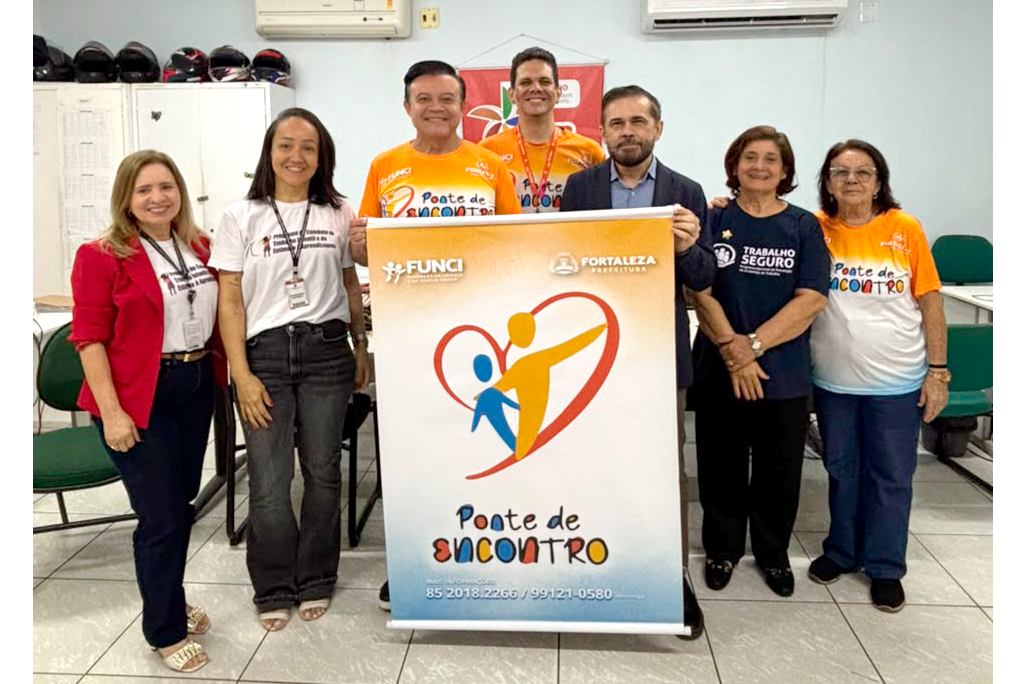 Sete pessoas adultas estão em pé em uma sala de escritório posando para a foto. Duas delas, no centro, seguram um banner vertical com a marca da FUNCI e da Prefeitura de Fortaleza e o título "Ponte de Encontro" acima da ilustração de um coração. A intenção é divulgar e registrar o lançamento ou a atuação de um programa social e institucional focado no acolhimento e apoio comunitário.