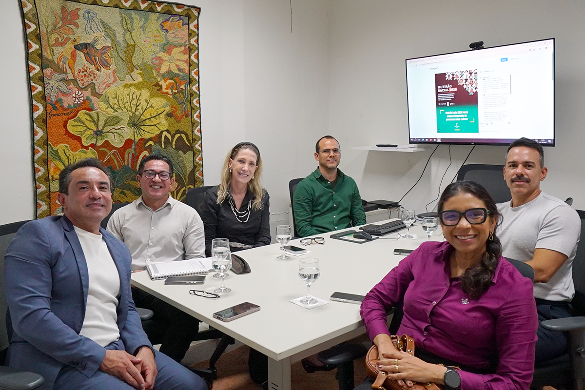 Imagem fotográfica. Seis pessoas estão reunidas em uma sala de reuniões, sentadas ao redor de uma mesa branca, olhando sorridentes para a câmera. Ao fundo, há uma tapeçaria colorida na parede e uma televisão que exibe um cartaz digital com o título "MUTIRÃO RACIAL 2026". A cena transmite uma mensagem de colaboração, trabalho em equipe e alinhamento institucional para a organização do evento.