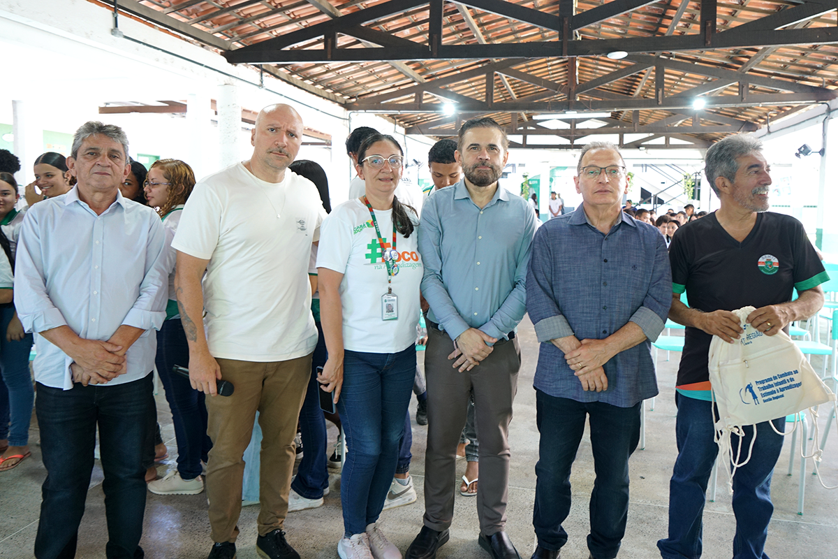 Fotografia institucional apresentando seis adultos alinhados lado a lado no pátio coberto de uma escola, onde se veem estudantes ao fundo. Eles posam para a foto em tom solene e amigável, com as mãos cruzadas à frente ou segurando objetos, como um microfone e uma sacola de projeto social. A intenção da imagem é registrar a presença e a união de autoridades e educadores durante uma ação comunitária ou evento escolar.