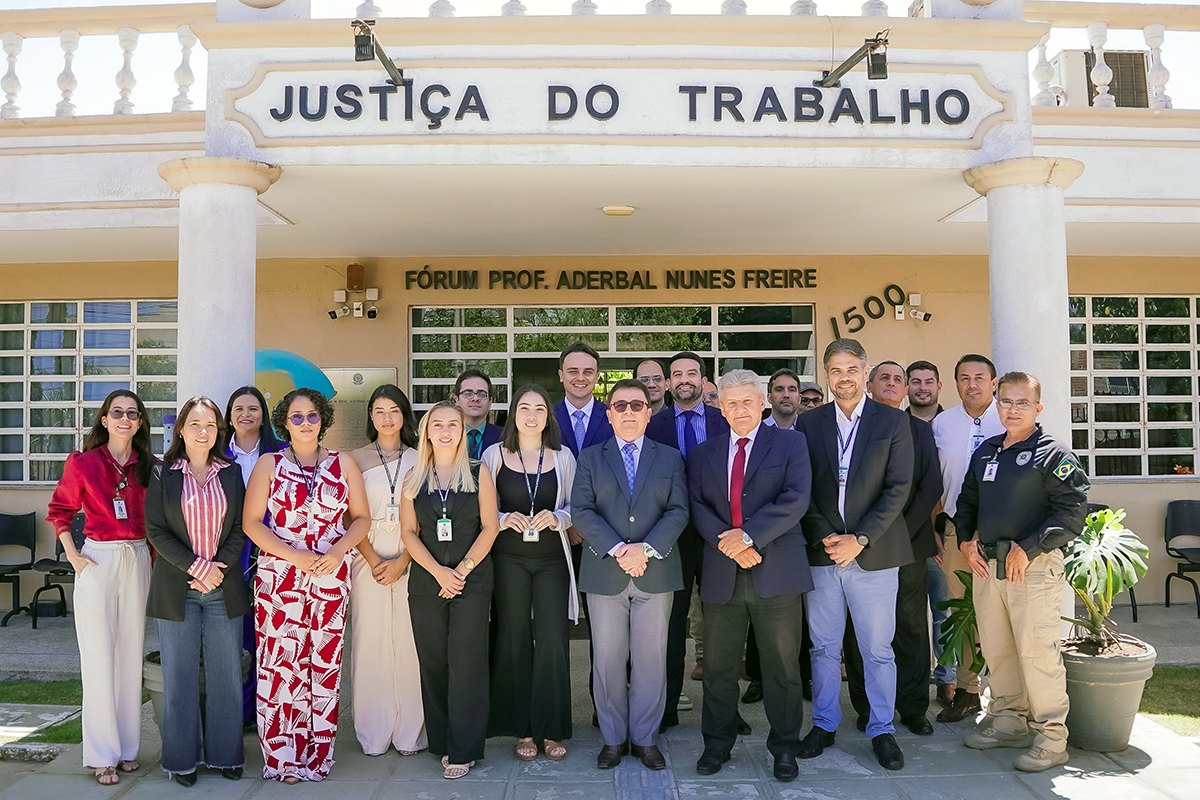 Grupo de cerca de 18 pessoas pose lado a lado em frente ao prédio do Fórum Prof. Aderbal Nunes Freire, da Justiça do Trabalho. Ao fundo, lê-se "JUSTIÇA DO TRABALHO" em destaque sobre a fachada de tom bege com detalhes brancos. Homens e mulheres vestem trajes esporte fino, sociais e uniformes, sorrindo para a câmera em um dia ensolarado. No canto direito, há um vaso com uma planta costela-de-adão.
