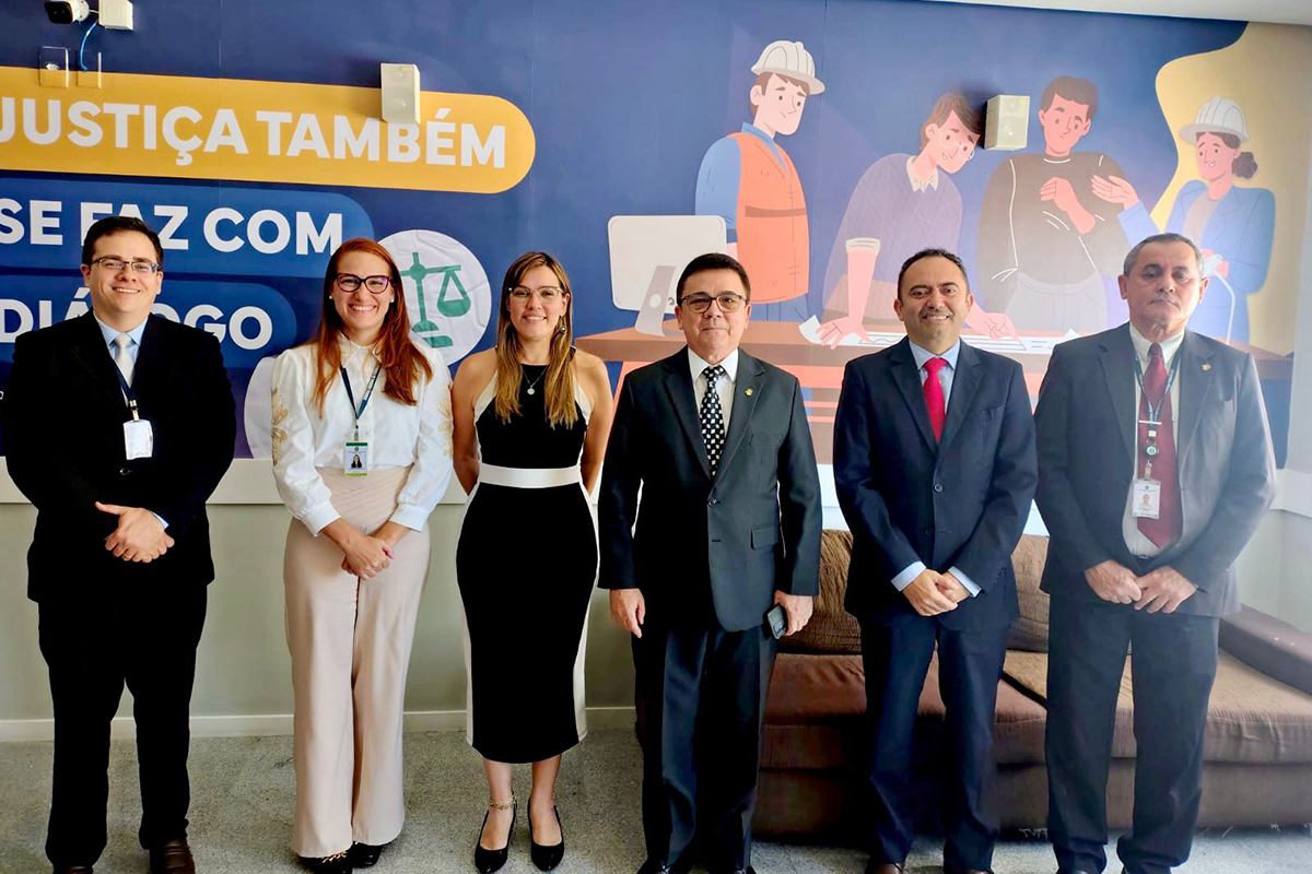 Seis profissionais, sendo quatro homens de terno e duas mulheres em trajes sociais, posam alinhados em um ambiente interno. Ao fundo, um painel azul exibe a frase "JUSTIÇA TAMBÉM SE FAZ COM DIÁLOGO" junto a ilustrações de trabalhadores e uma balança. Todos sorriem para a câmera, transmitindo uma postura formal, cordial e de cooperação institucional.
