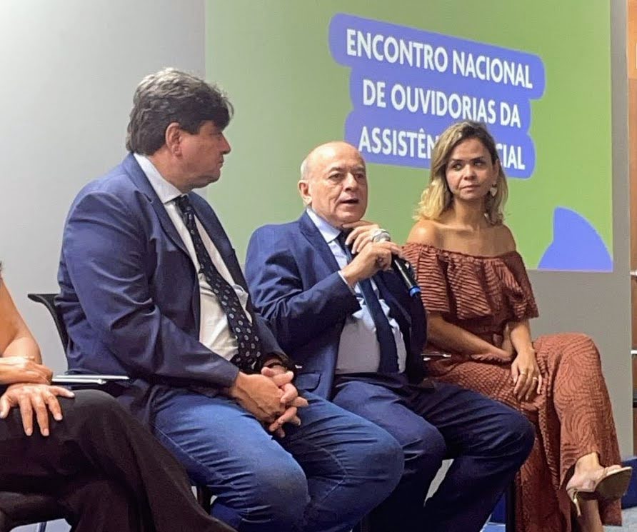 A imagem mostra um painel de evento com três pessoas sentadas em cadeiras estofadas escuras. O foco está no palestrante central, um homem careca de meia-idade, vestindo um terno azul-marinho e gravata clara. Ele segura um microfone com a mão direita e parece estar falando ou fazendo uma pausa na fala.  À sua esquerda (do ponto de vista do observador), há um homem corpulento, de cabelos castanhos escuros e revoltosos, vestindo um blazer azul-marinho sobre uma camisa e gravata escuras, com calça jeans azul. Ele olha para o palestrante central, com uma expressão séria e atenta.  À direita do palestrante (do ponto de vista do observador), está uma mulher jovem de pele morena, com cabelos loiros cacheados na altura dos ombros. Ela veste um vestido ou blusa off-shoulder (decote ombro a ombro) marrom, com um tecido texturizado de estampa sutil. Ela olha diretamente para a frente, também com uma expressão séria.  Ao fundo, em uma tela ou painel projetado, há um fundo verde e azul com texto em português que começa com as palavras em destaque em azul claro: ENCONTRO NACIONAL DE OUVIDORIAS DA ASSISTÊNCIA SOCIAL.  A iluminação é clara e focada no painel.