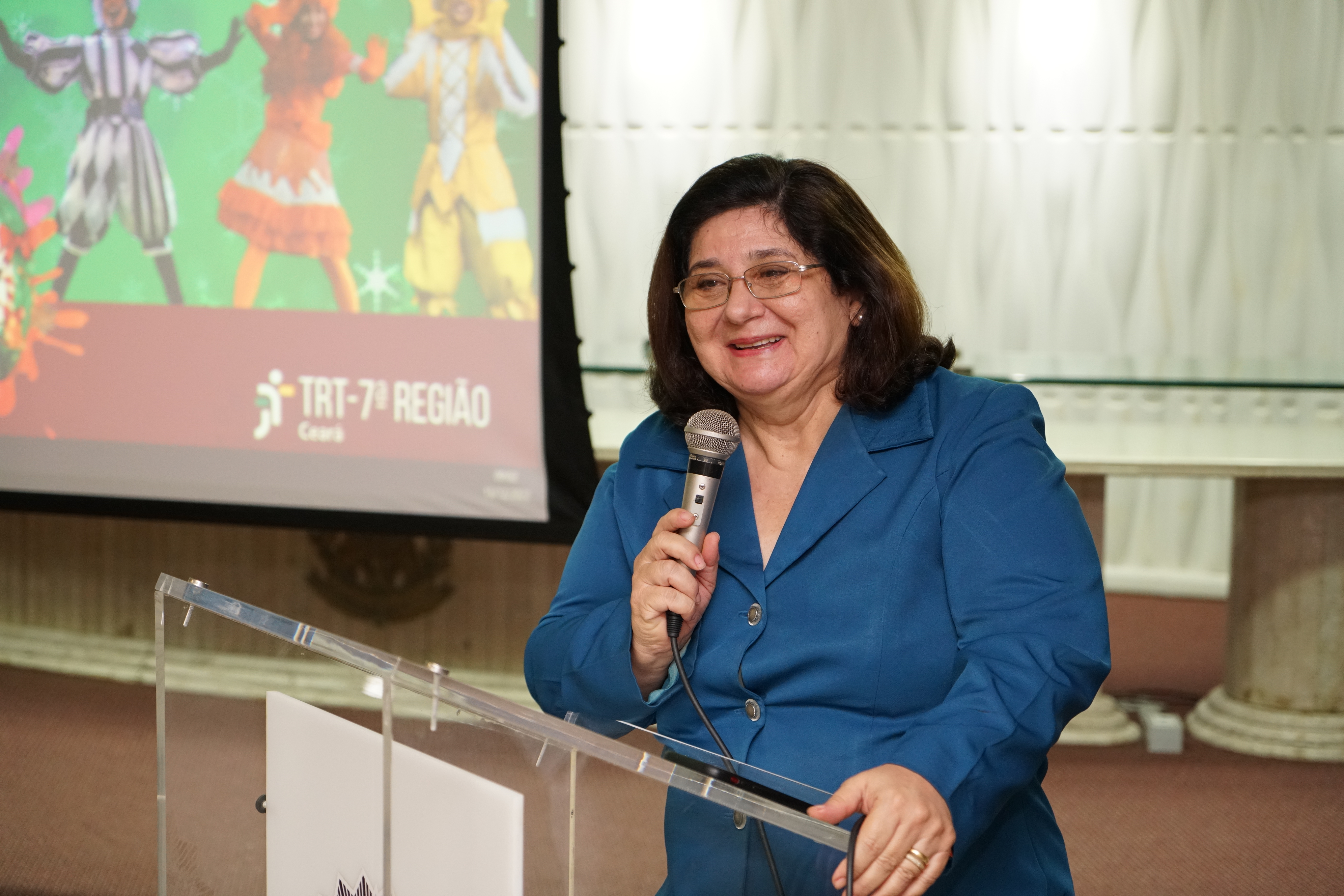 Desa. Regina Gláucia falando no microfone sorridente. A magistrada está no púlpito do auditório